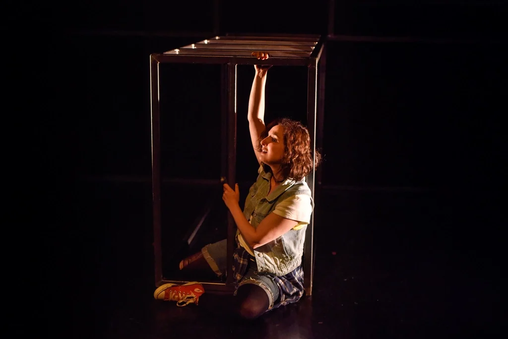 Actrice au sol, tenant une structure métallique avec une expression d’espoir.