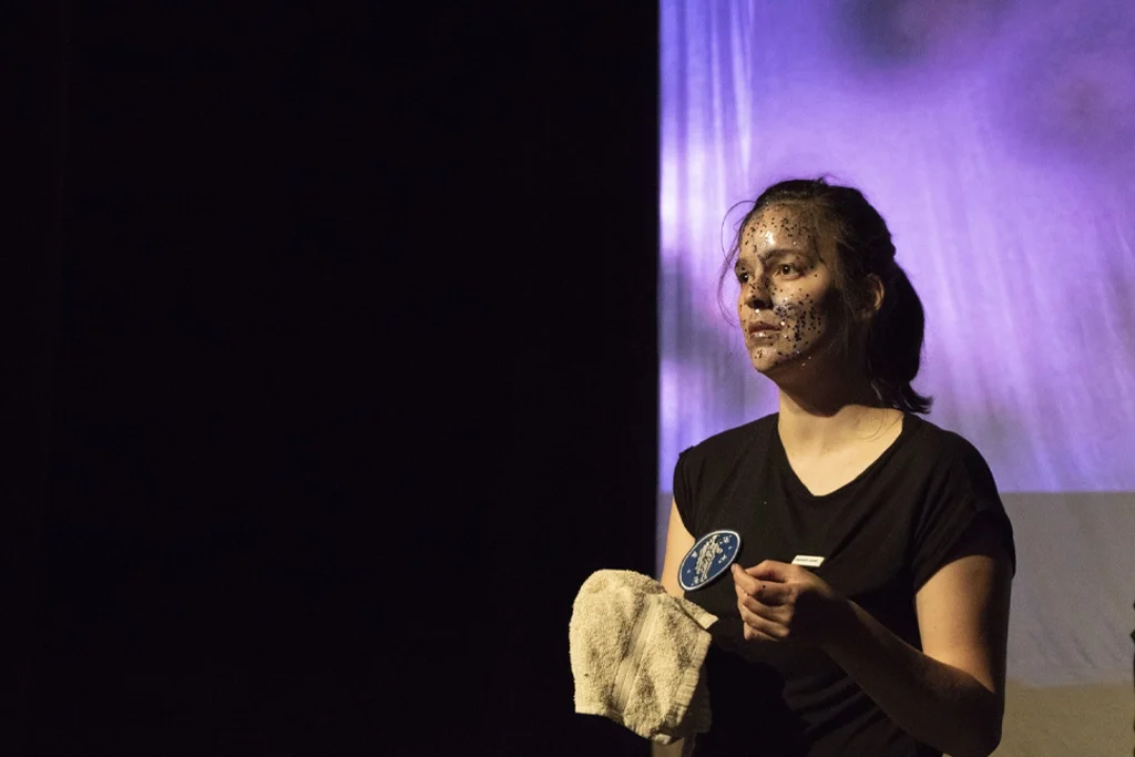 Une actrice avec un maquillage étoilé sur le visage, tenant un chiffon sur scène pendant la pièce « Planétarium ».