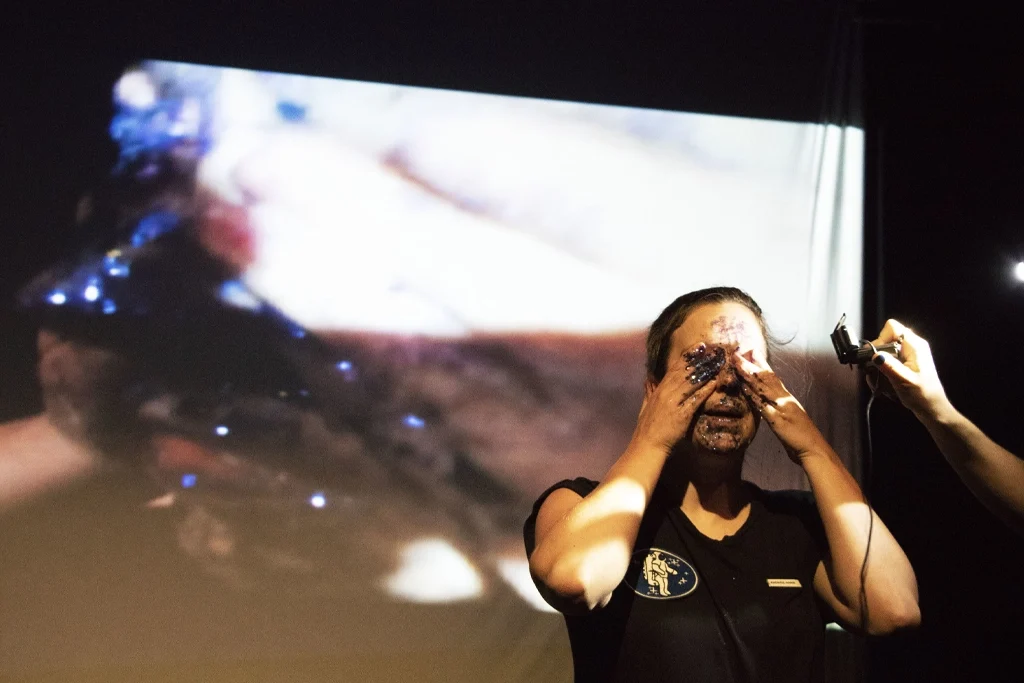 Actrice sur scène, les mains sur le visage, avec une projection visuelle de gros plan sur écran en arrière-plan lors de la pièce « Planétarium ».